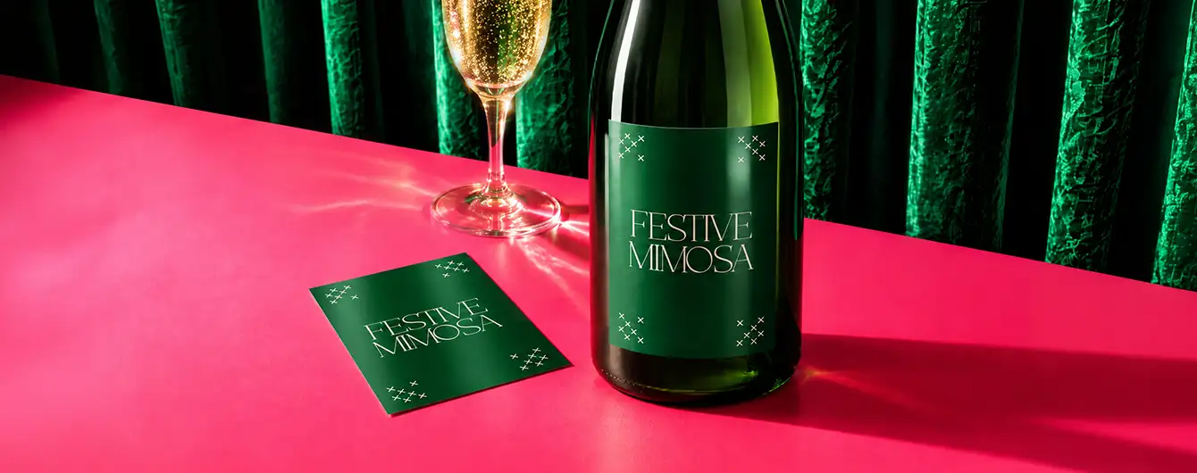 A green wine bottle with a matching green Bottle Label next to a matching green card, a glass of champagne, and a green curtain backdrop on a pink surface.