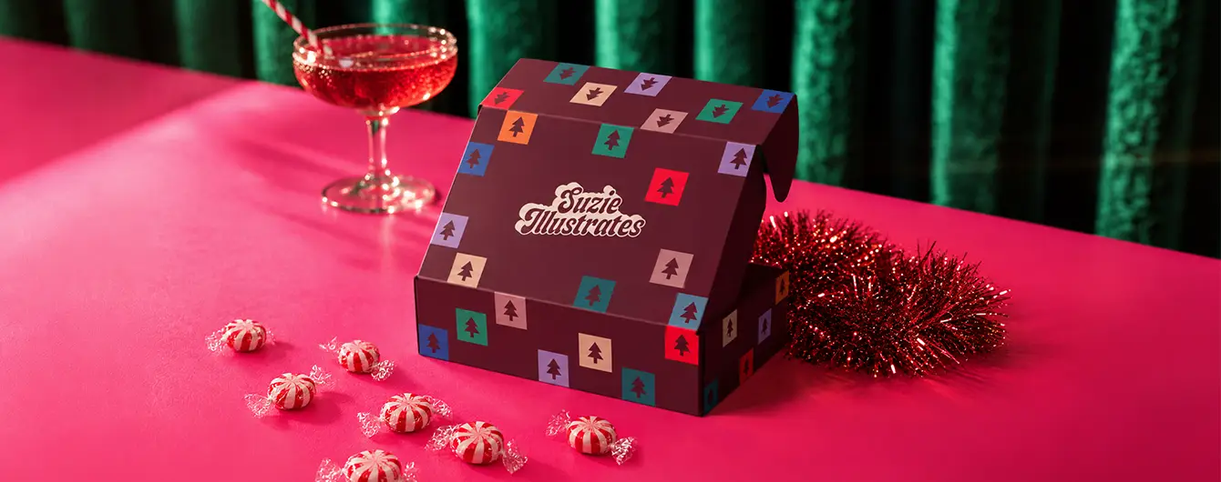 A dark burgundy patterned gift Box resting on a bright pink tabletop alongside red tinsel, wrapped peppermint candies, and a pink cocktail in a coupe glass.