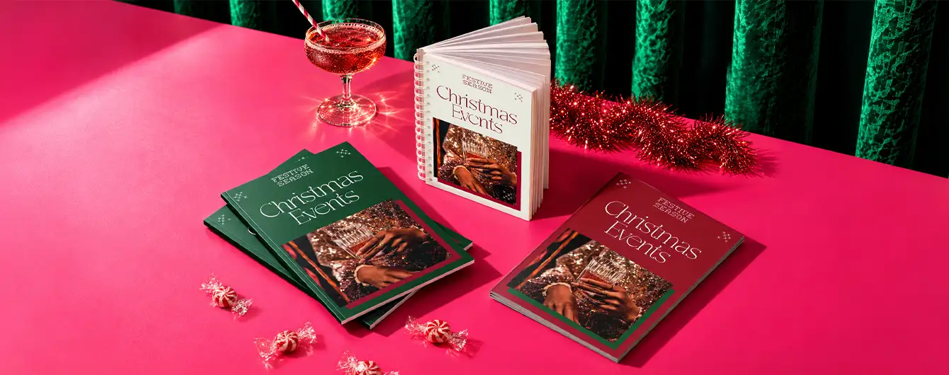 Three "Christmas Events" Booklets -one green, one burgundy, and one standing open in white—displayed on a pink surface alongside a cocktail glass, red garland, and wrapped candies.