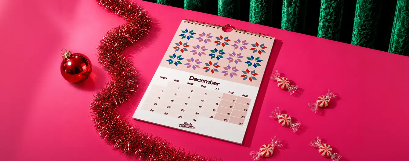 n open spiral-bound Calendar displaying the month of December on a bright pink surface, surrounded by red garland, a Christmas bauble, and scattered peppermint candies.