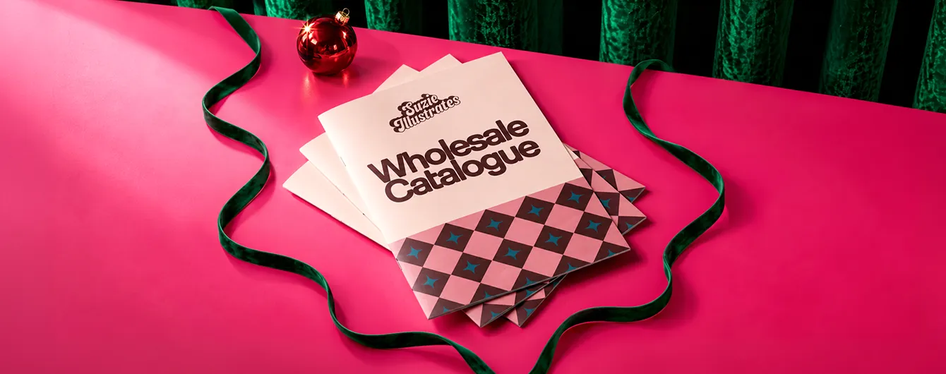 A stack of "Suzie Illustrates Wholesale Catalogue" booklets with a pink and brown diamond-patterned design, resting on a pink table with green ribbon and a red ornament