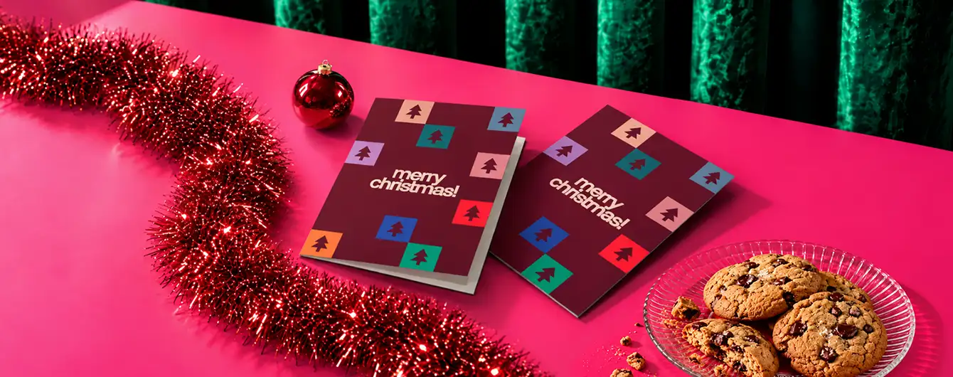 Two dark burgundy "Merry Christmas" Christmas Cards placed on a bright pink surface next to red tinsel, a red ornament, and a small plate of chocolate chip cookies