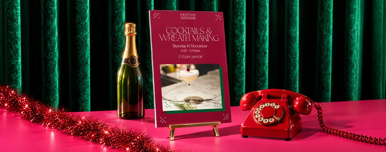 A tall maroon Display Board sign reading "Cocktails & Winter Sipping" displayed on a stand alongside a bottle of champagne, a red vintage rotary phone, and red garland on a pink surface