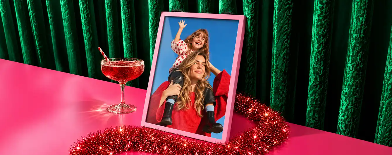 Framed Print in a pink frame showing a mother carrying a young child on her shoulders against a blue sky, displayed on a bright pink table alongside a festive cocktail glass and red tinsel garland against a green velvet curtain background.