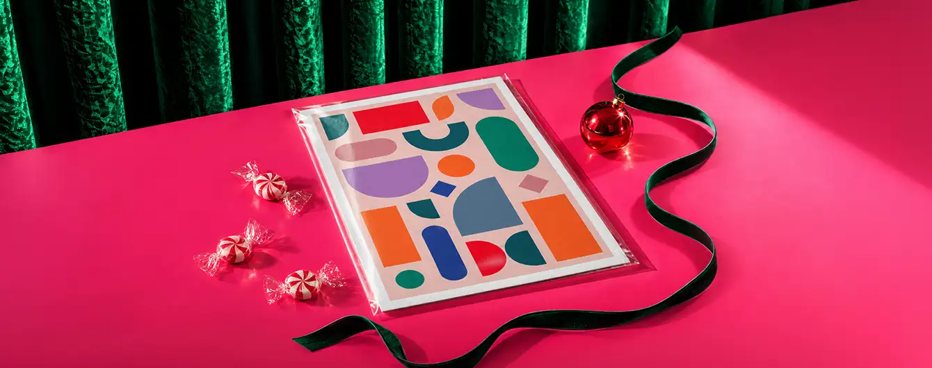 A Giclee art print featuring a colourful abstract geometric design, displayed on a pink surface with green ribbon, a red ornament, and peppermint candie