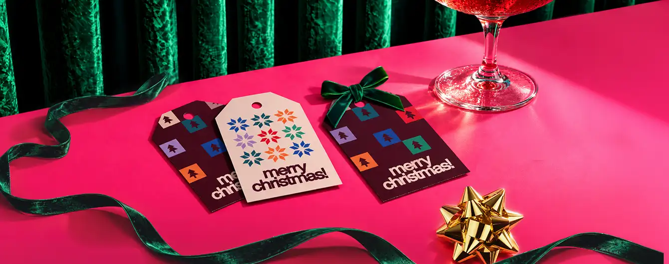 hree Christmas Gift Tags featuring colourful geometric patterns and "merry christmas!" text displayed on a pink surface alongside a green velvet ribbon, a gold bow, and a cocktail glass