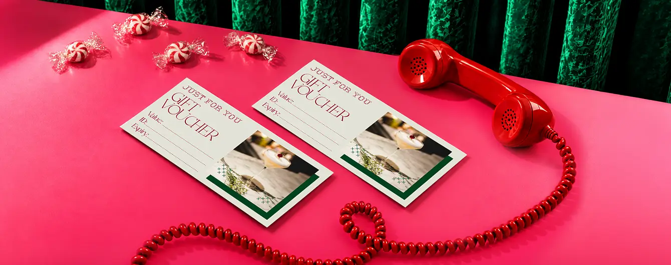 Two cream-coloured "Gift Voucher" cards with red text and a cocktail photo, displayed on a pink table next to a red rotary phone and wrapped candies