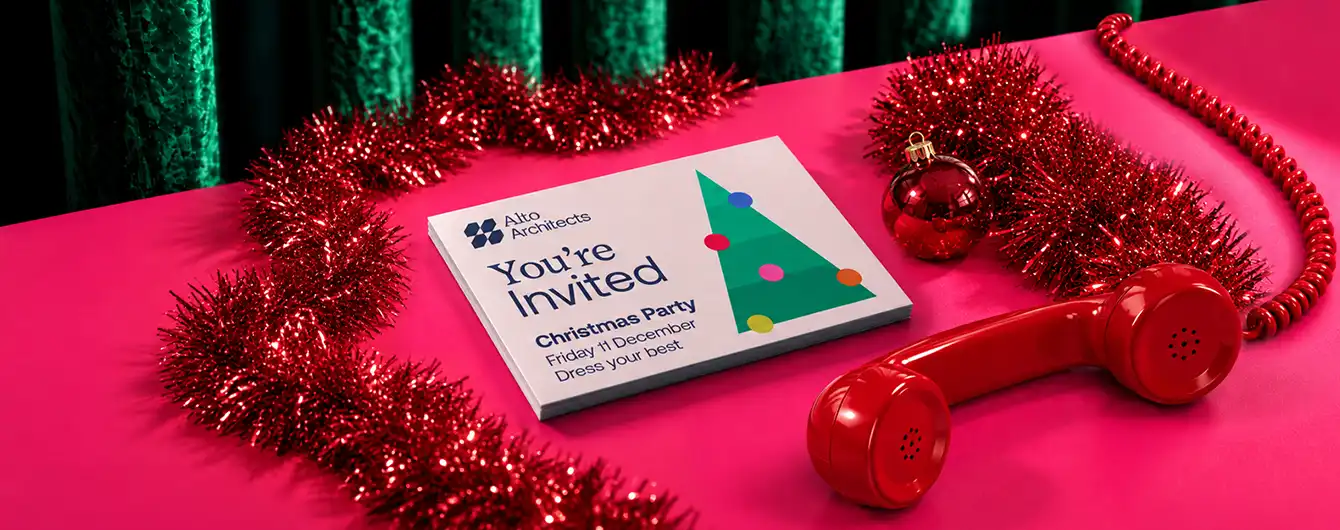 Stack of Christmas party Invitations featuring a green Christmas tree illustration, displayed on a pink surface with red tinsel garland, a red ornament, and a vintage red telephone handset.
