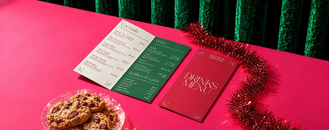 An open folded Menu with white and green panels next to a red "Drinks Menu" booklet, displayed on a pink surface with a plate of cookies and red tinsel garland.