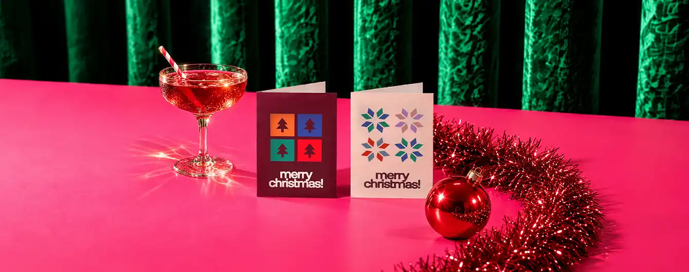 Two standing "Merry Christmas" Mini Greeting Cards displayed on a pink table next to a festive cocktail, a red ornament, and red garland