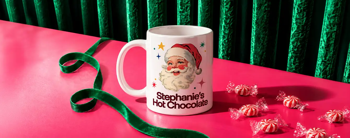 A white ceramic photo Mug printed with a vintage Santa Claus illustration and the text "Stephanie's Hot Chocolate," set on a bright pink surface with a coiled green ribbon, wrapped peppermint candies, and a green velvet curtain background