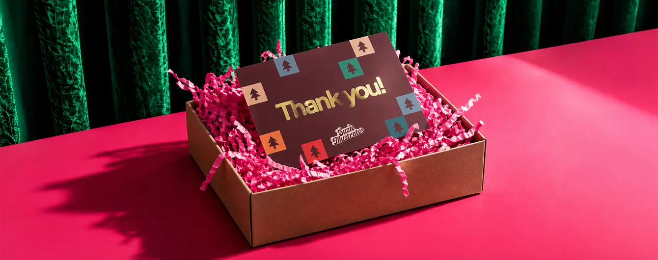 A dark burgundy "Thank you!" Packaging Insert Card sitting inside a cardboard box filled with pink crinkle paper on a bright pink surface.