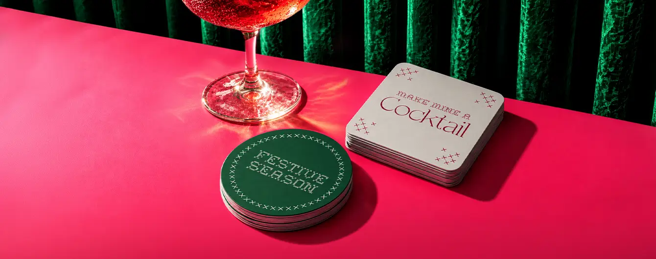 A stack of white square beverage Coasters and a stack of round green drink coasters displayed on a pink surface next to a glass of wine