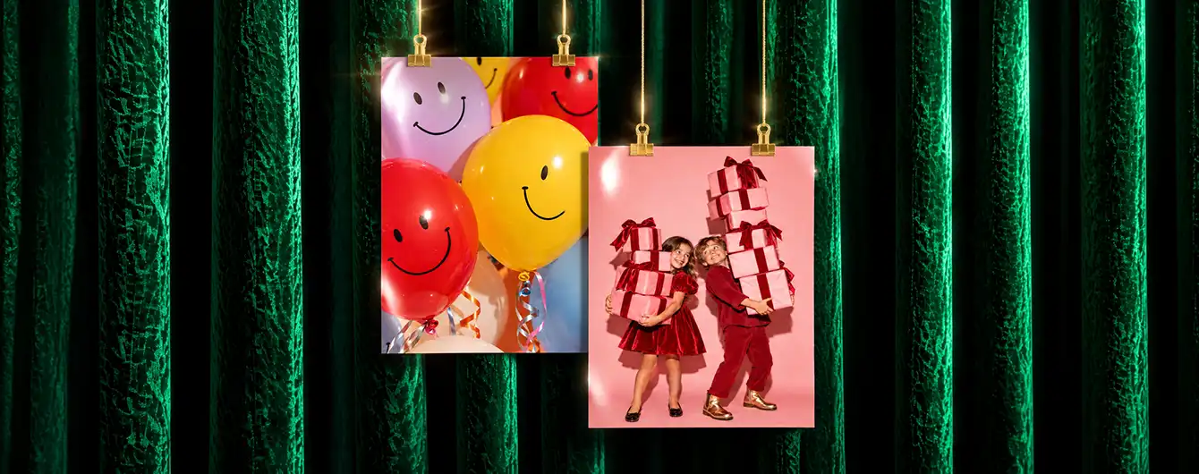 Two Photography Prints clipped to brass cords against a green velvet curtain background—one featuring colorful balloons with smiley faces, and the other showing two children in red holiday outfits holding stacked gifts