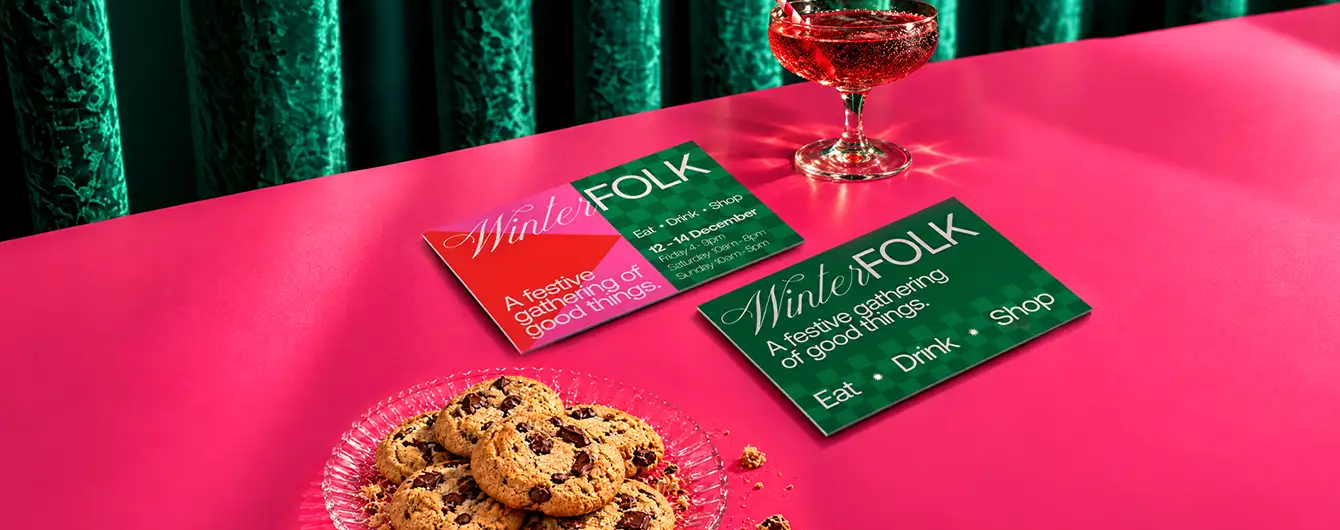 Two colourful "Winter FOLK" promo Postcards laid out on a pink surface next to a glass of cocktail and a plate of chocolate chip cookies