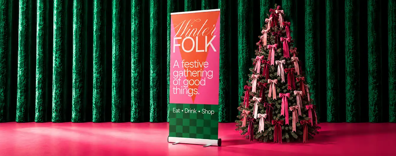 : A tall "Winter FOLK" Roller Banner stand next to a Christmas tree decorated with pink and red bows against a green velvet curtain background