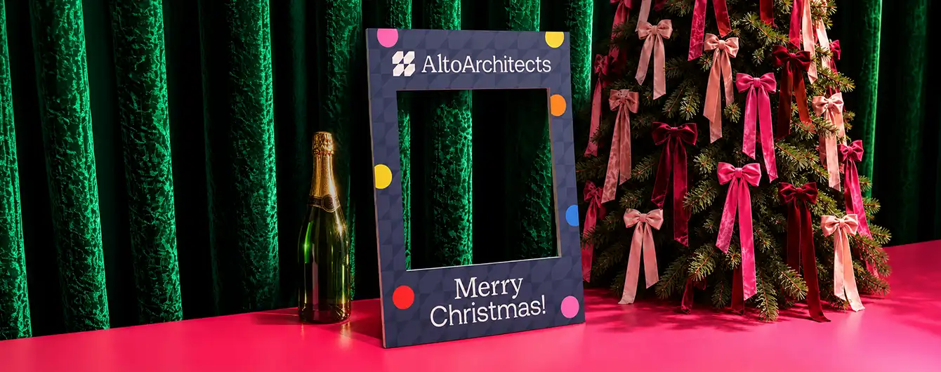 A dark blue photo booth frame decorated with colorful dots and "Merry Christmas!" text, standing on a pink surface next to a bottle of champagne and a Christmas tree adorned with bows.