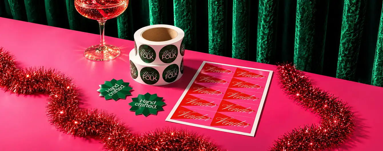 A sheet of red Christmas Stickers, with Sticker Rolls and green starburst Die Cut Stickers on a pink surface with red tinsel garland and a glass of rosé wine.