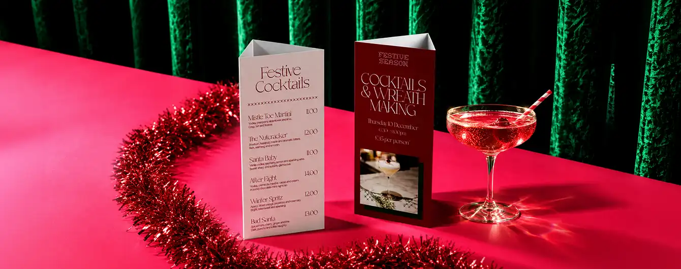 Two standing Table Talker Menus—one white and one maroon—displayed on a pink surface next to a festive cocktail and red tinsel garland