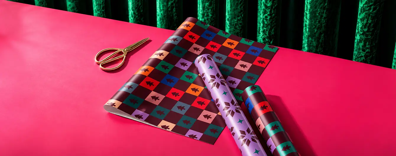 Sheet and roll format Christmas Wrapping Paper featuring colourful geometric patterns displayed on a pink surface next to a pair of gold scissors.