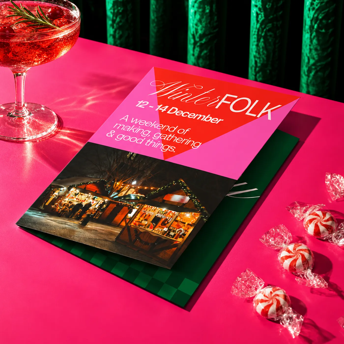 Christmas Folded Leaflet showcasing "Winter Folk" market details and imagery, displayed with festive sweets and seasonal décor on a vibrant pink tabletop.